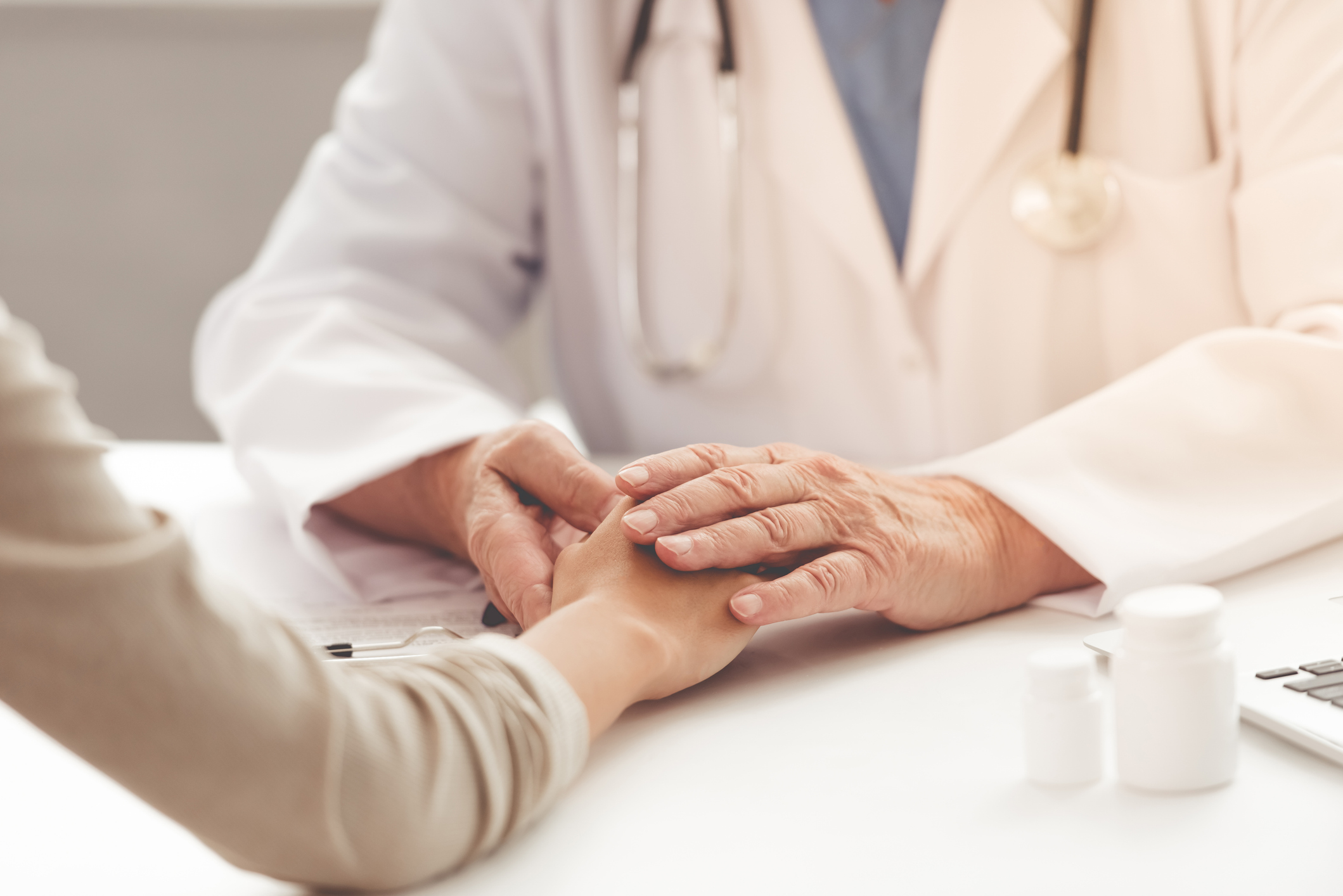Cropped image of old doctor in white medical coat consulting his patient and holding her hand while sitting in his office